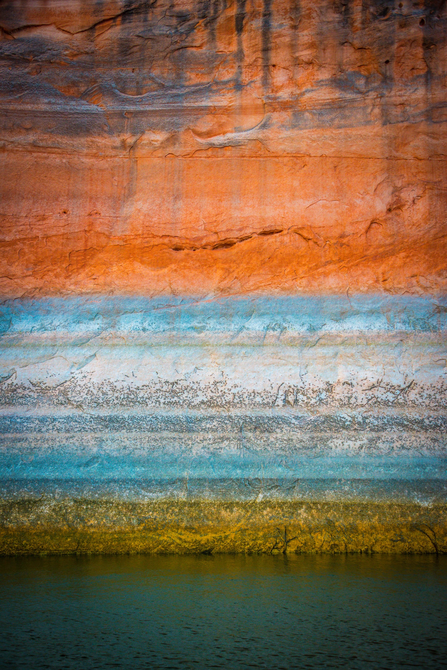 A beautiful gradient of green, blue, white, grey, red, orange, and black of the Glen canyon walls, sitting over the waters of Lake Powell, Utah. The colors tell the story of changes in water levels, minerals, and chemistry in the lake over time. As if time painted its story on the walls.